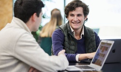 Two young men are having a conversation at a table with laptops in a modern environment.