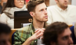 A young man with a green plaid shirt is listening attentively in a classroom setting.