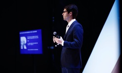 A speaker in a suit is presenting on stage with a microphone.