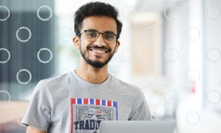 A smiling young man with glasses is sitting in front of a laptop in a modern workspace.
