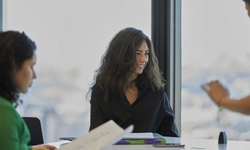 A woman with long, wavy hair smiles during a meeting in a modern office space.