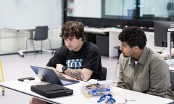Two students are working together at a table with a laptop and electronics components in a modern classroom.