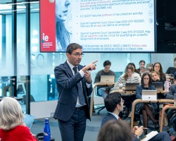 A man gestures while speaking in front of an audience during a presentation.