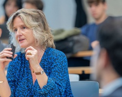 A woman with curly hair is speaking in front of an audience in a classroom setting.