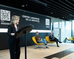 A woman speaks at a podium in a modern event space featuring a QR code backdrop and flags.