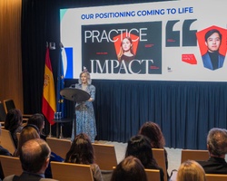 A presentation event with a speaker addressing an audience while visuals are displayed on a screen.