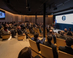 A large audience attentively listening to a speaker on stage in a modern conference room.