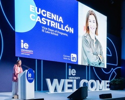 A woman stands at a podium in front of a large screen displaying a welcome presentation at IE University.