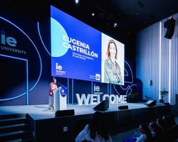 A speaker presenting at an event with a large screen displaying their name and university logo.