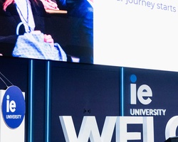 A woman stands at a podium welcoming attendees to a university event.