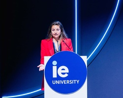 A speaker in a red outfit stands at a podium with the IE University logo.