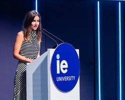 A woman is speaking at a podium with the logo of IE University in a modern setting.