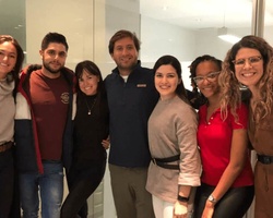 A group of seven people smiling and posing together indoors, with a sign in the background indicating no smoking.