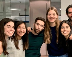 A group of six young adults smiling and posing together in an office environment.