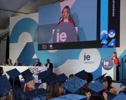 A graduation ceremony at IE University with students in blue gowns and a speaker on stage.