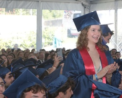 A young woman in a graduation cap and gown stands smiling and applauding among fellow graduates at a commencement ceremony.