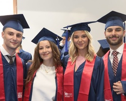 Four graduates in blue and red regalia smiling at a graduation ceremony