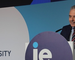 A man in a suit speaking at a podium with the IE University logo on it.