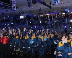 Graduation ceremony with students in blue and yellow gowns tossing their caps in the air, indoors with spectators watching from balconies.
