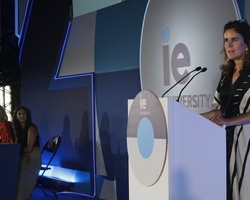 A woman giving a speech at a podium with a large 'IE University' backdrop and the slogan 'Ready for the Future' during an event.