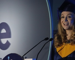 A female graduate giving a speech at a podium with a logo of 'ie University' during a graduation ceremony.