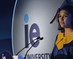 A female graduate in a blue and yellow gown giving a speech at a university podium.