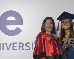 A young graduate in a blue cap and gown smiles as she stands next to a woman in a red stole, both posing in front of a banner of IE University.