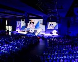 A large audience at a conference with a speaker on stage and multiple screens displaying the event.