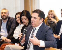 A man in a business suit speaking into a microphone at a seminar, surrounded by an attentive audience.