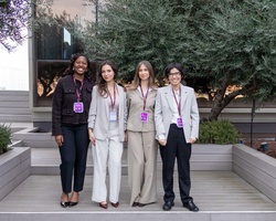 Four women are standing together on an elevated platform surrounded by greenery and a modern building.