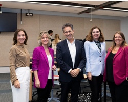 Five professionals, three women and two men, smiling in a conference room with chairs and tables in the background.