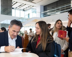 A man signs a book at a table while conversing with a young woman in a crowded event space.