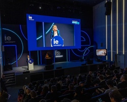 A speaker presents on stage at a large event with an audience in attendance.