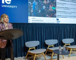 A woman speaks at a podium in a conference room with a banner of IE University and the European flag in the background, and an empty seating area to her right.