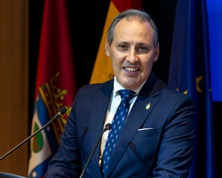 A man in a suit is speaking at a podium with flags in the background.