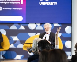 Two men sitting in front of an audience at a university event, with one speaking and the other listening.