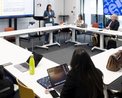 A woman stands presenting to a group of eight people seated around a conference table in a modern office.