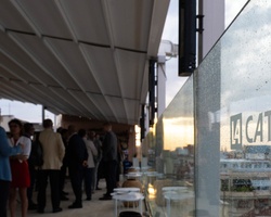 The image depicts a social gathering on a rooftop terrace with a view, featuring people chatting and glass barriers with reflections.