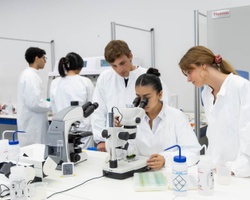 A group of students in white lab coats are engaged in a science experiment involving a microscope.