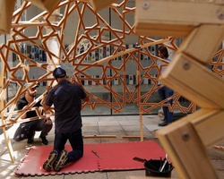 Two people are assembling a large geometric wooden structure outdoors.