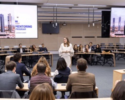 A woman stands presenting at the front of a conference room filled with seated participants, with two large screens displaying 'MENTORING PROGRAM'.