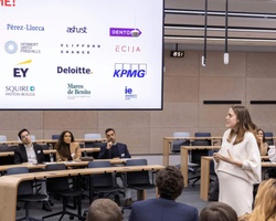 A woman stands speaking at the front of a lecture hall filled with seated attendees, with several professional service firm logos displayed behind her.