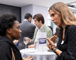Two women engage in conversation at a professional networking event, with other attendees in the background.