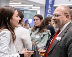 A young woman and an older man engaging in a conversation at a networking event, with other attendees in the background.