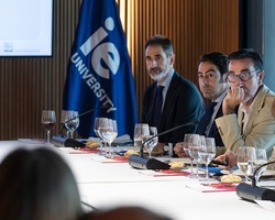 A group of people engaged in discussion at a conference table with a presentation screen in the background.
