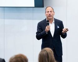 A man in a suit is giving a presentation to an audience in a modern conference room.