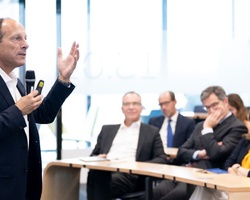 A man is speaking at a podium in a business seminar while a group of professionals listens attentively.