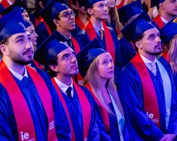 A diverse group of graduates wearing caps and gowns, looking up with anticipation during a ceremony.