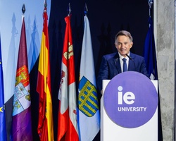 A speaker stands at a podium with various flags in the background during an event for IE University.