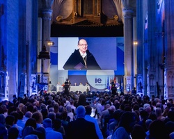 A large audience attending a presentation in a grand hall illuminated in blue light.